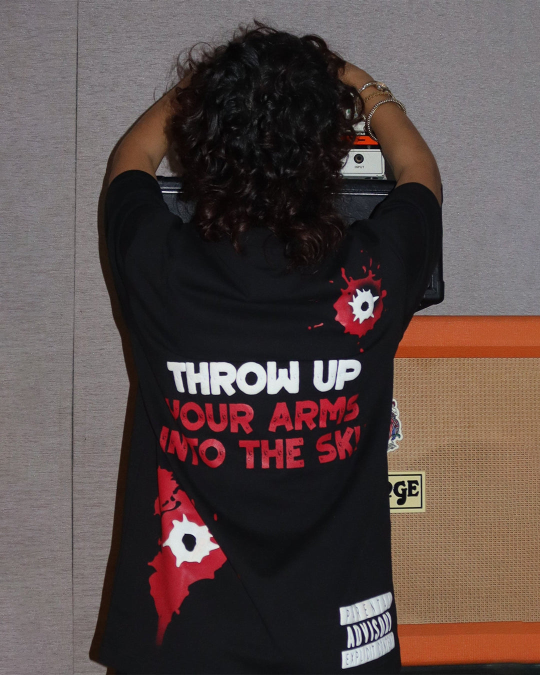woman wearing black tshirt with three head/hair outlines in the front , in the colour yellow green and orange ,and throw up your arms into the sky written on the back