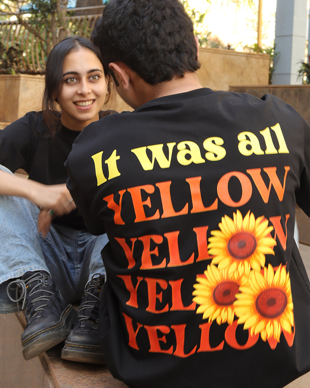 Woman sitting wearing a black t-shirt, With a bright orange sun in the front at the pocket side and a man sitting with her with it was all yellow written on the back