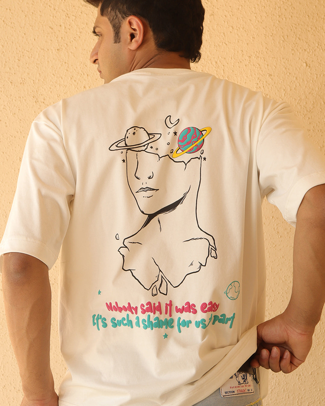 Man standing wearing a white t-shirt, With a art in the front at the pocket side and art and "nobody said it was easy , its such a shame for us to part" written on the back