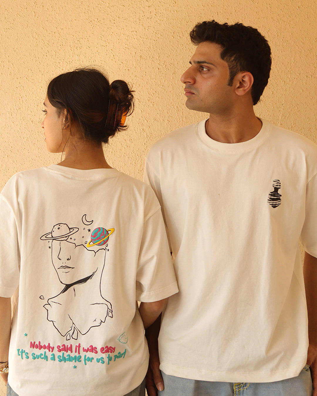 Woman and Man standing wearing a white t-shirt, With a art in the front at the pocket side and art and "nobody said it was easy , its such a shame for us to part" written on the back