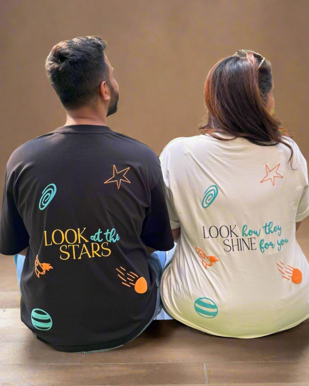 Man and Woman sitting wearing a black and White t-shirt, with "Look at the stars" and "Look how they shine for you" written in the back