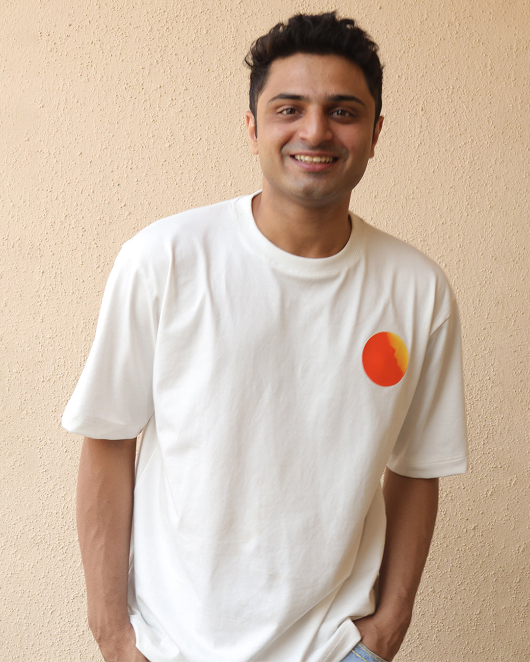 Man standing wearing a White t-shirt, With a bright orange sun in the front at the pocket side and it was all yellow written in the back