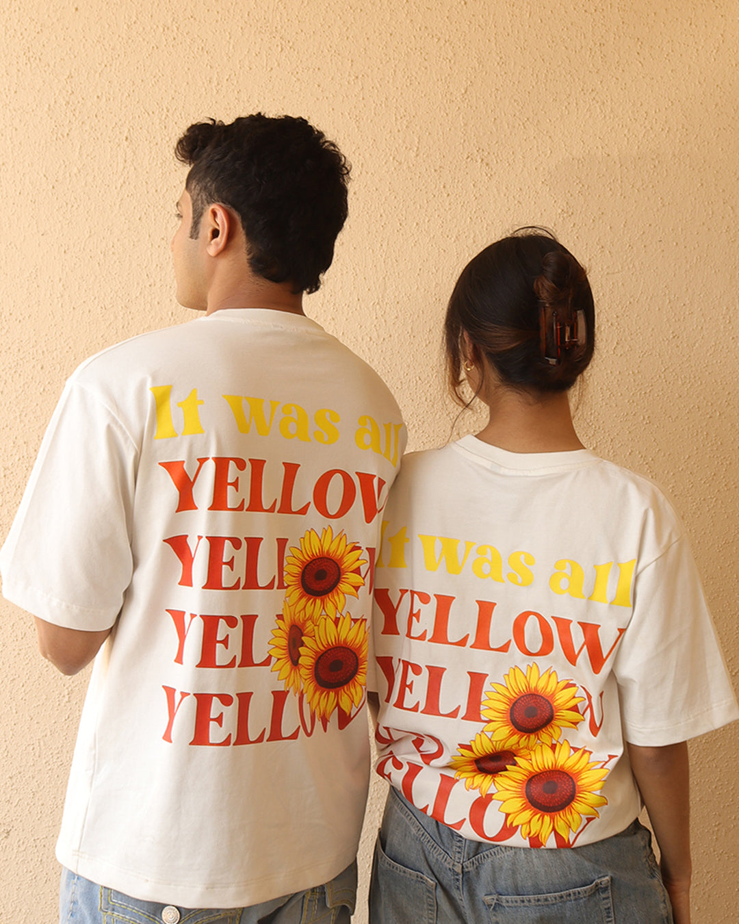 Woman and a man standing wearing a white t-shirt, With a bright orange sun in the front at the pocket side and it was all yellow written in the back