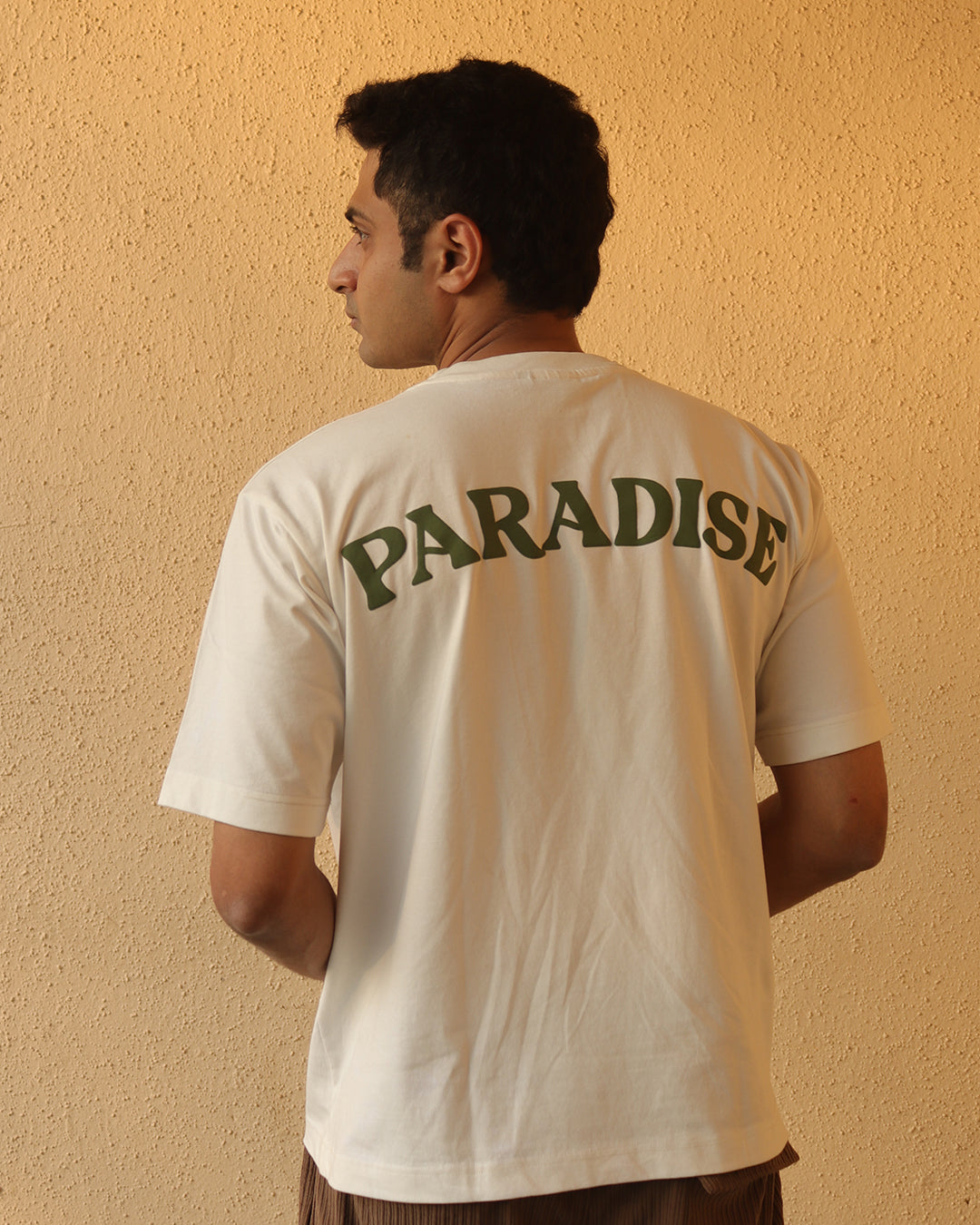 Man standing Wearing white t-shirt and Palm tree on the pocket side and paradise written on the back