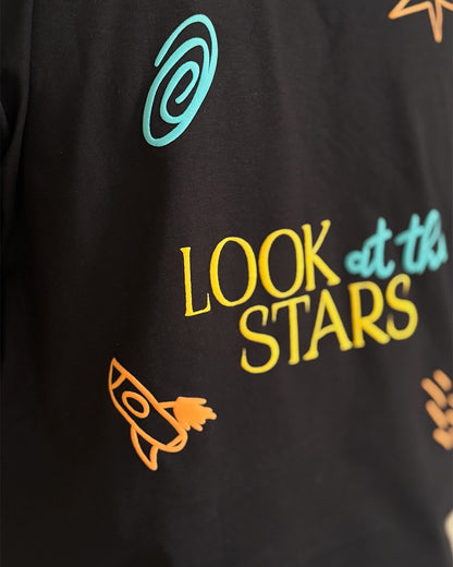 Man Standing and wearing a black and black t-shirt, with "Look at the stars" written in the back