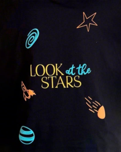 Man Standing and wearing a black and black t-shirt, with "Look at the stars" written in the back